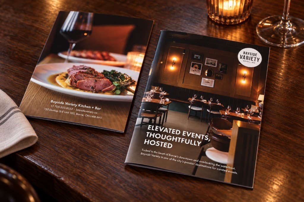 Two Bayside Variety brochures on a dark wood table: one showing a plated dish, the other a dining-room photograph and a short heading about hosted events.