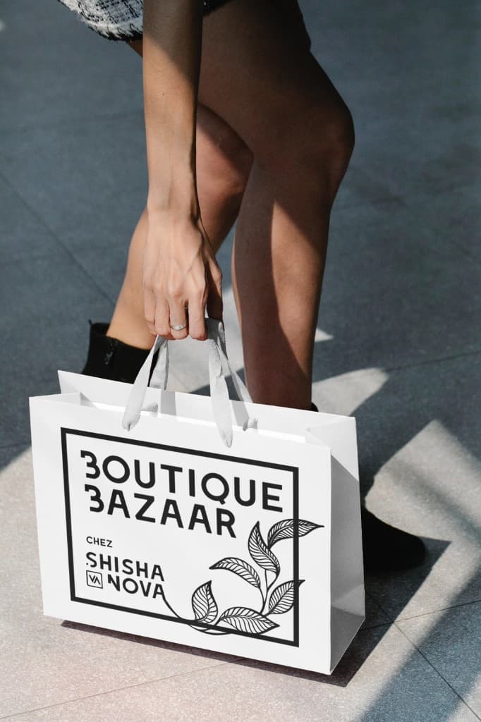 A person carrying a white paper shopping bag printed with the black Boutique Bazaar chez Shisha Nova logo and leaf illustration.