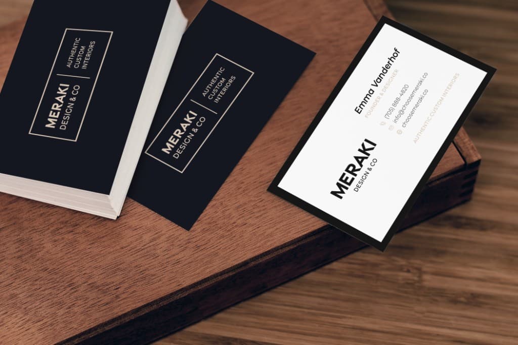 Meraki Design Co. business cards on a wood surface: a stack of black cards with a gold-framed wordmark, and a white contact card.