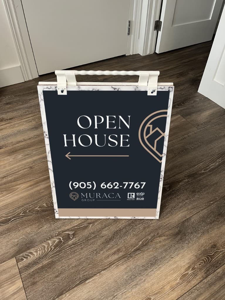 A Muraca Group open-house sandwich board on a wood floor, navy with a gold house-and-pin mark and a phone number.