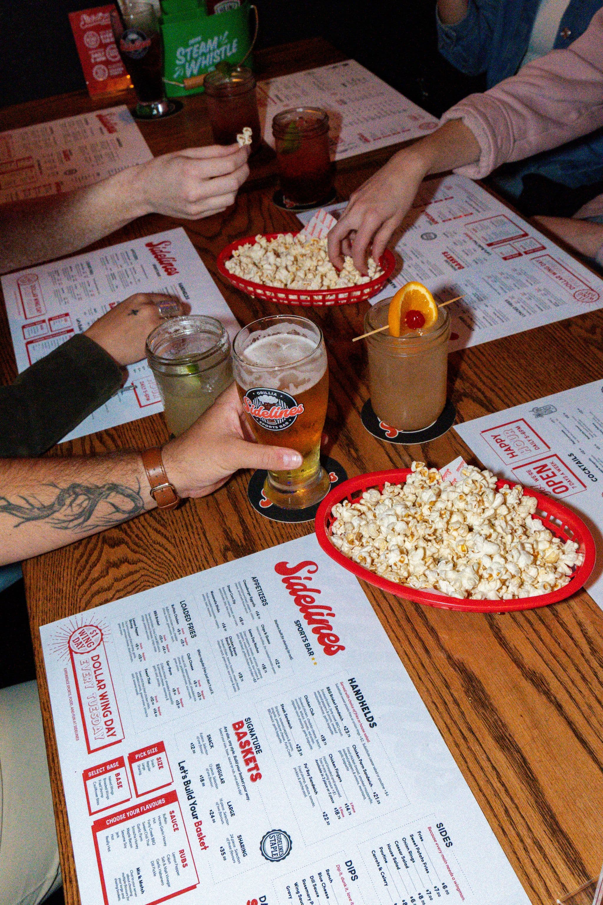 Guests at a wooden bar table with beer, cocktails, and baskets of popcorn on top of Sidelines Sports Bar paper menus with red section headings.