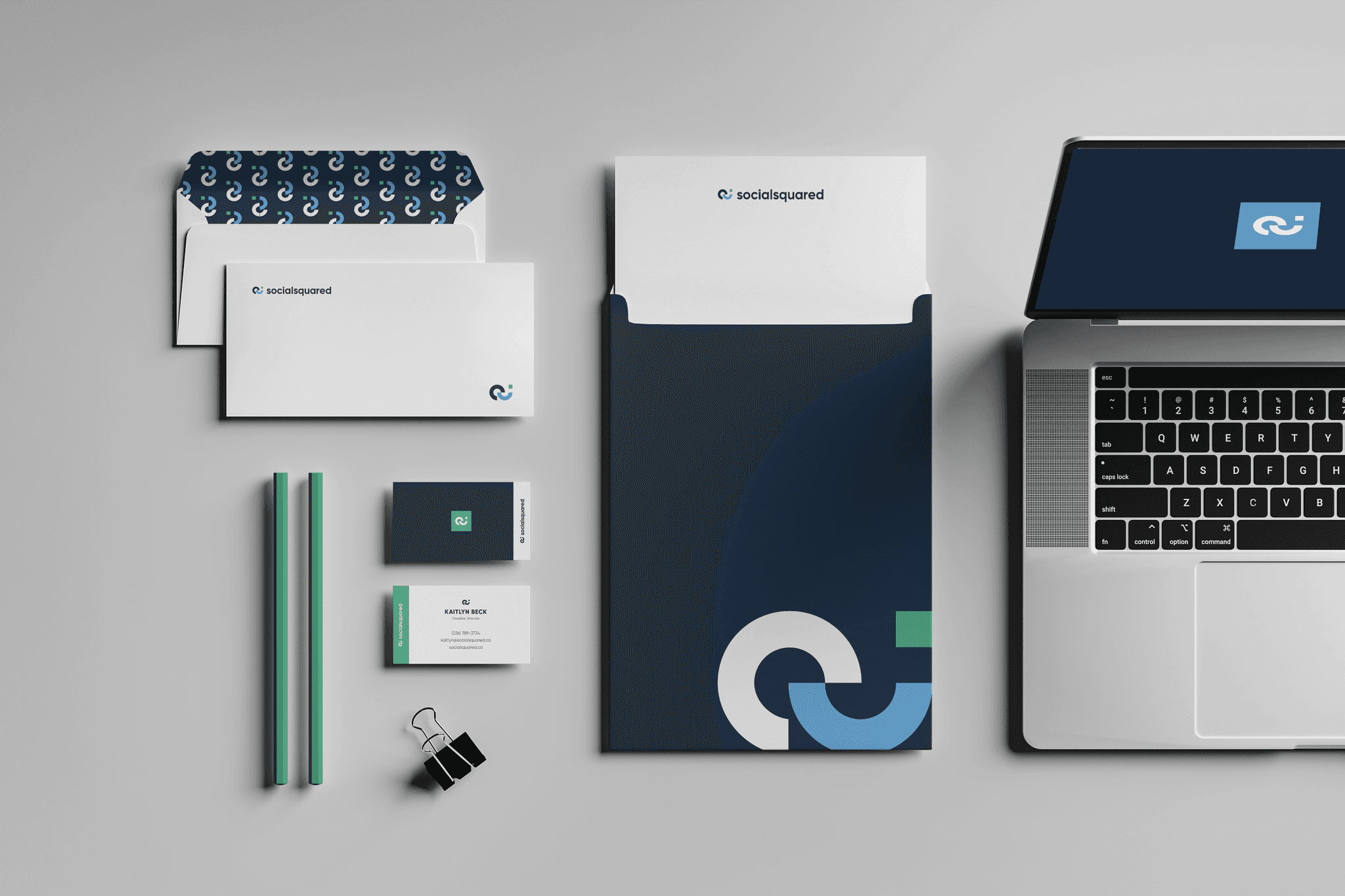 SocialSquared stationery laid out flat: patterned envelope liner, letterhead, navy presentation folder with the interlocking S mark, business cards with green accents, and a laptop showing the logo.