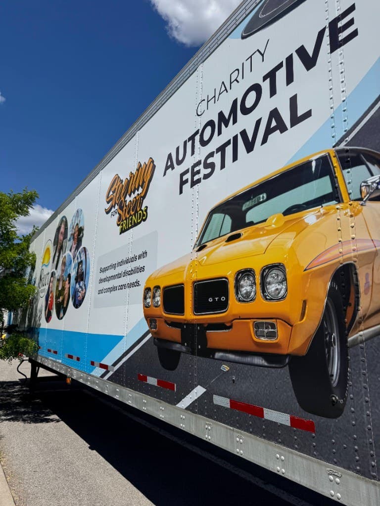 Close view of the Steer Friends trailer wrap: a yellow classic car, circular portraits, and large type for the charity automotive festival.