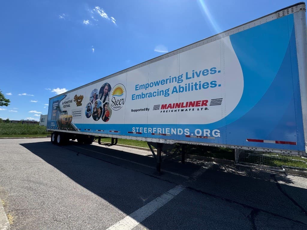 A Steer Friends trailer parked on asphalt under a clear sky, wrapped in white and blue with circular photos, the wordmark, Empowering Lives. Embracing Abilities., and steerfriends.org.