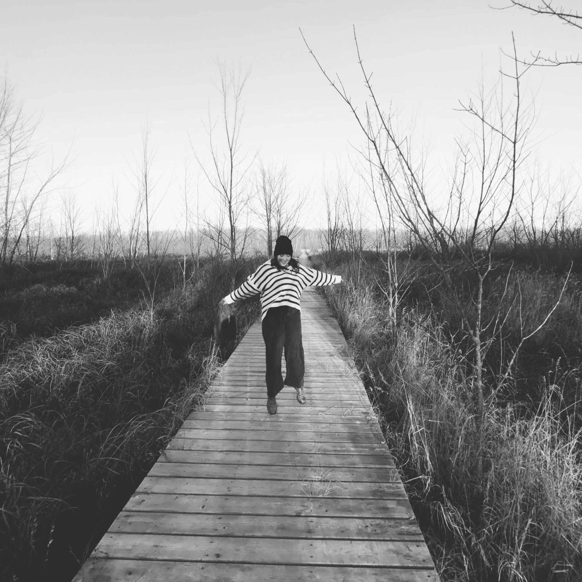 Kaitlyn Beck in a striped sweater and toque walking along a wooden boardwalk through tall grass and bare trees, arms outstretched. Black and white photograph.