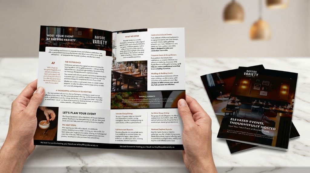 Hands holding an open Bayside Variety event brochure beside a stack of dark covers on a pale table.