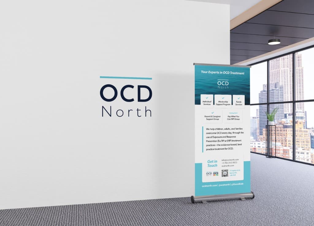 An office lobby with the OCD North wordmark on a white wall and a roll-up banner showing service tiles and contact details.
