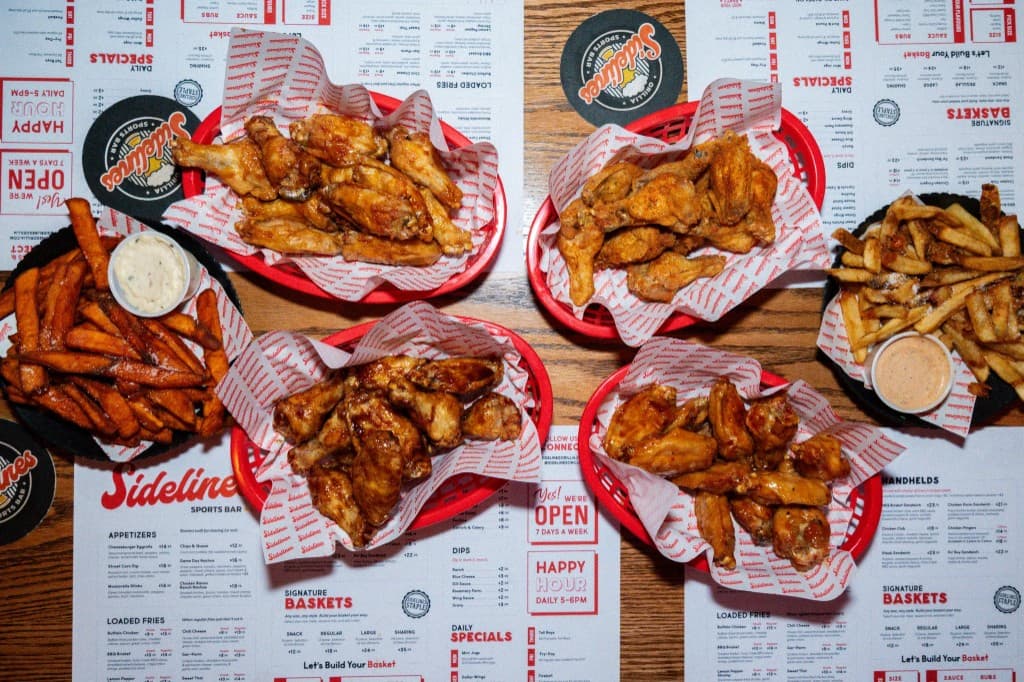 Overhead view of red baskets of chicken wings and fries on a wooden table covered with Sidelines paper menus and black logo coasters.