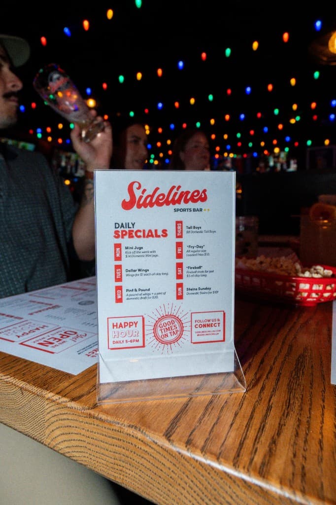 Acrylic table tent on a bar listing Sidelines daily specials by weekday, with Happy Hour, Good Times On Tap, and Follow Us badges at the bottom, string lights blurred behind.