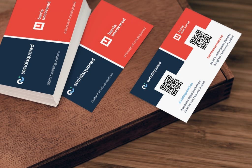 A stack of dual-branded cards on a wooden tray, navy and red faces up, beside one card turned to show its two QR codes and the taglines for each brand.
