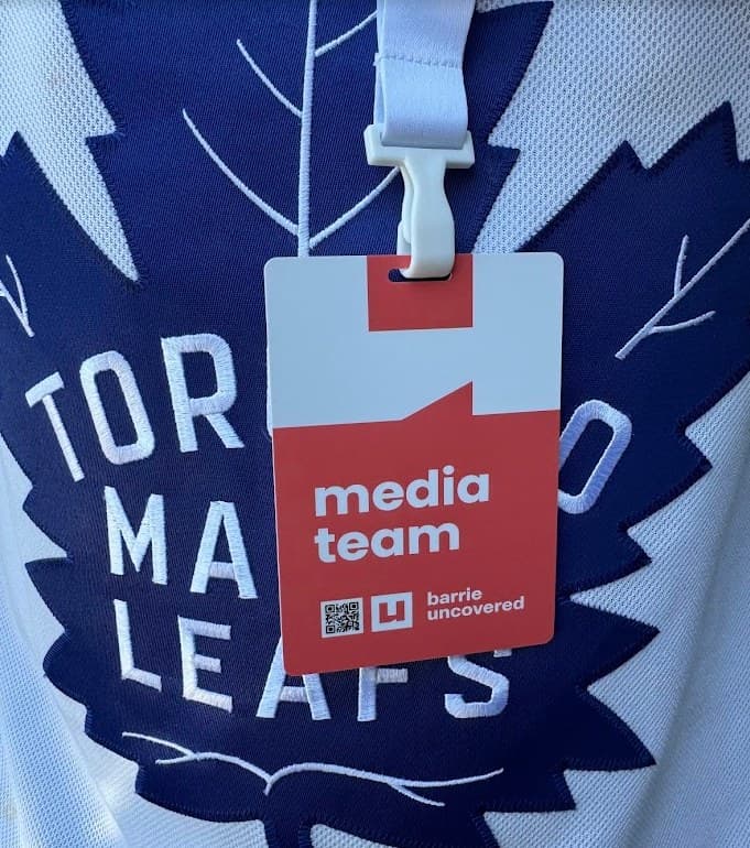 A Barrie Uncovered media-team badge on a white lanyard, hanging against a white hockey jersey with a blue maple leaf.