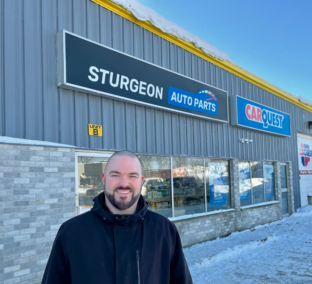 A man in a black jacket smiling in front of the Sturgeon Auto Parts storefront in winter, with the new black Sturgeon Auto Parts sign and a Carquest sign above the windows.