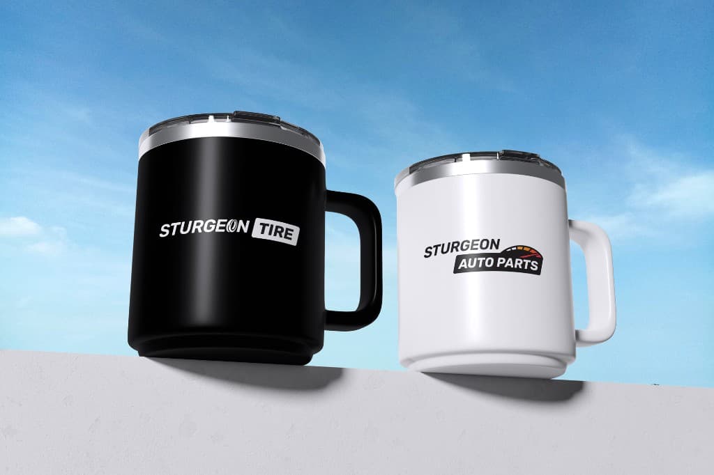 A black Sturgeon Tire mug beside a white Sturgeon Auto Parts mug on a pale ledge against a blue sky, presented as a mockup.