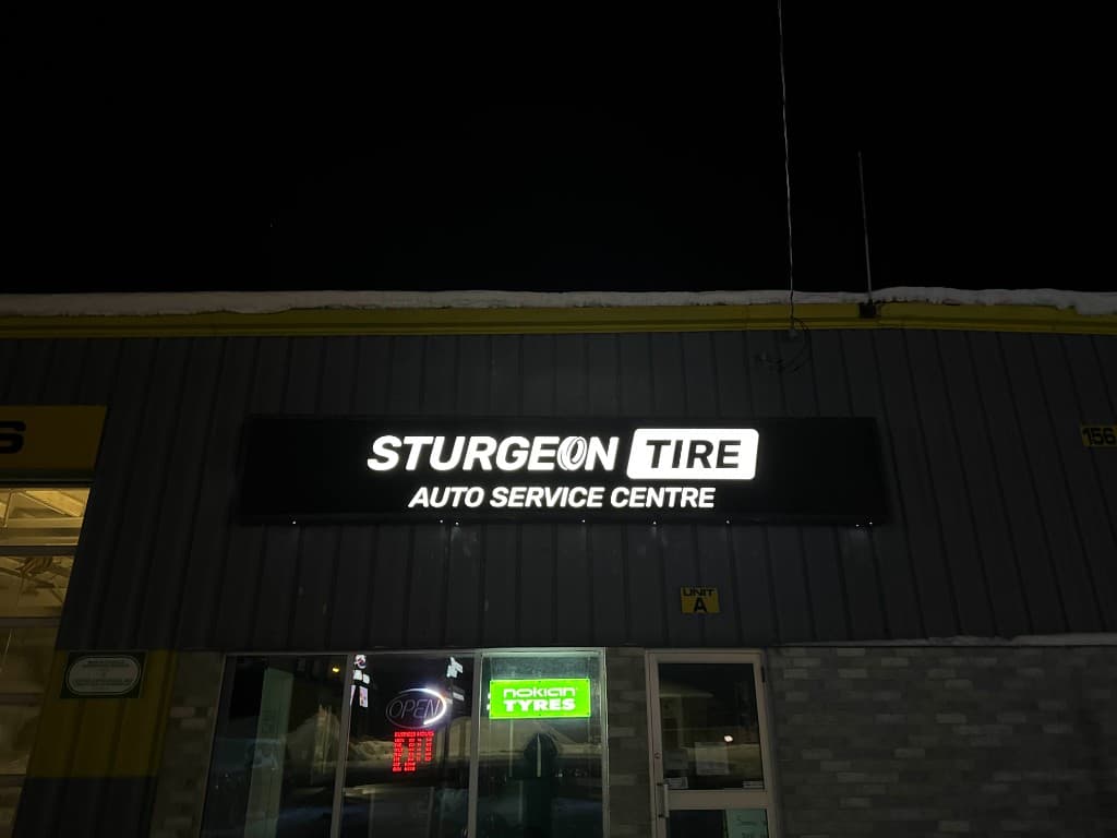 The illuminated Sturgeon Tire Auto Service Centre sign at night on a grey metal building, with the tire-shaped O in Sturgeon and TIRE set in a white rounded box.