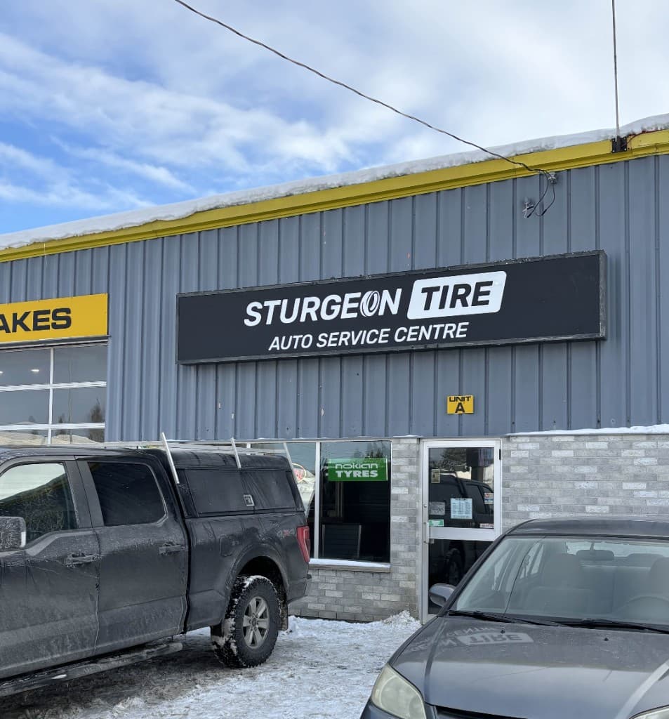 Daytime view of the Sturgeon Tire Auto Service Centre sign on a grey building with a yellow roofline, a pickup truck and a car parked in front.