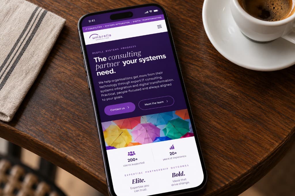 A phone on a dark wood table beside a coffee cup, showing the Umbrella Consulting homepage with a purple hero and a band of colourful umbrellas.